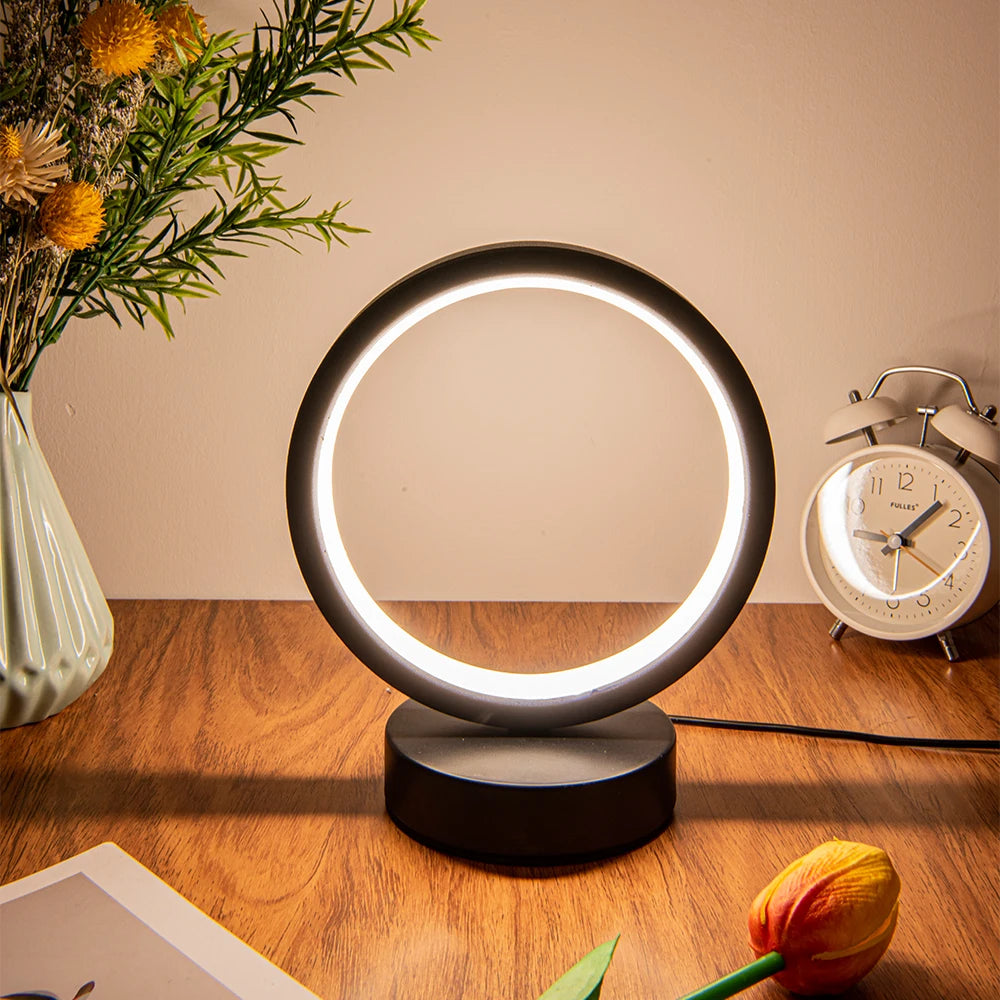 Glow & Drift Eclipse Minimalist Halo Lamp casting a warm white ambient glow on a wooden desk with a flower vase and a clock