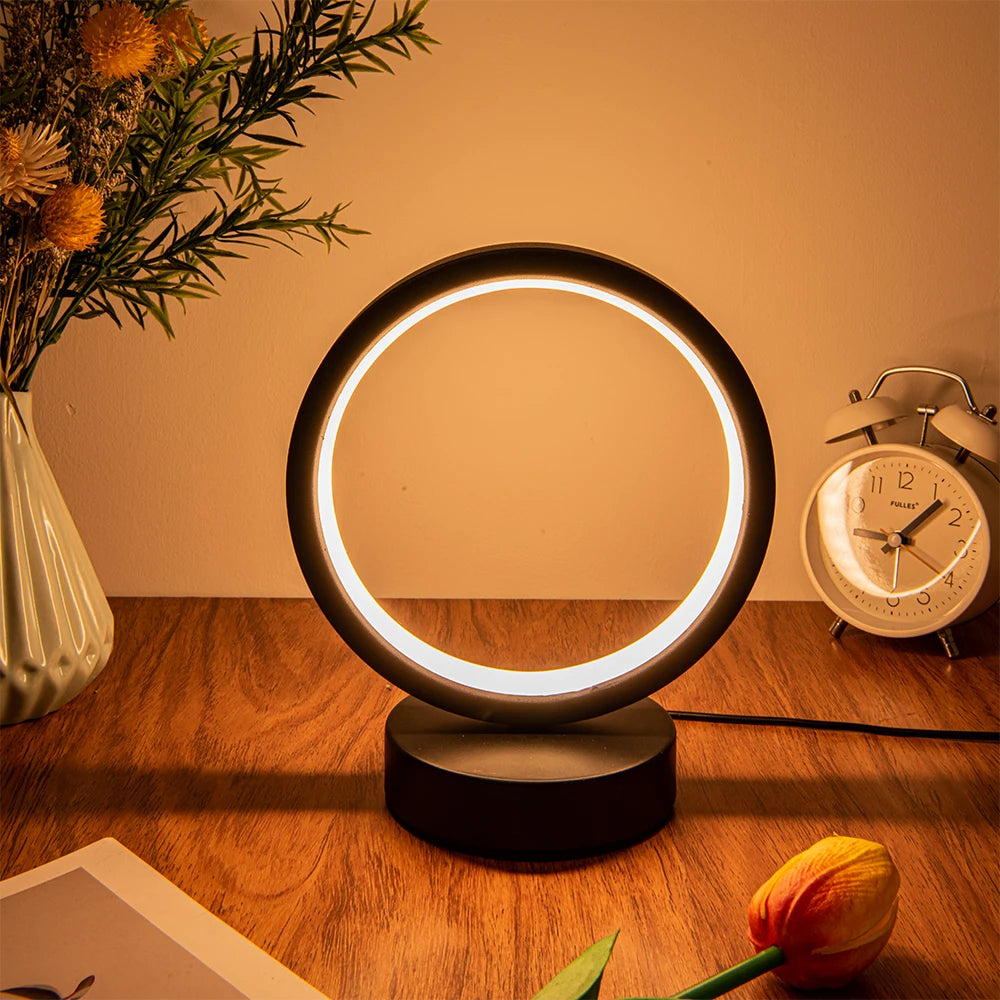 Glow & Drift Eclipse Minimalist Halo Lamp casting a warm white ambient glow on a wooden desk with a flower vase and a clock