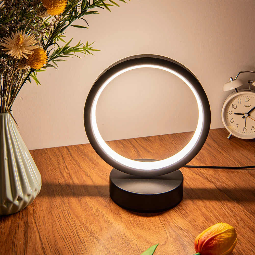 Glow & Drift Eclipse Minimalist Halo Lamp casting a warm white ambient glow on a wooden desk with a flower vase and a clock