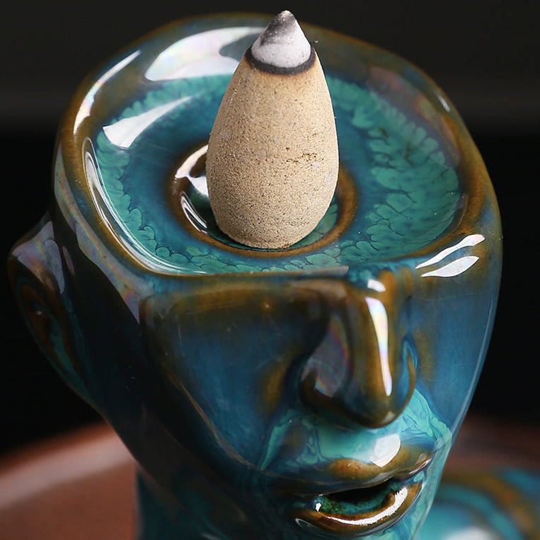 Extreme close-up of the top of a glazed teal ceramic bust incense burner, showing a lit backflow incense cone perfectly positioned in the specially designed holding cavity to enable the downward smoke effect.