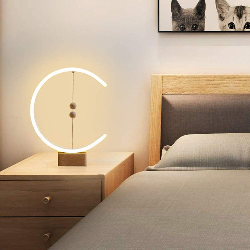 The Crescent magnetic balance lamp emitting a warm ambient glow on a wooden bedside table in a cozy, minimalist bedroom setting.