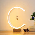 A close-up lifestyle shot of the crescent magnetic LED desk lamp glowing warmly on a light wood table next to decorative pinecones and books.