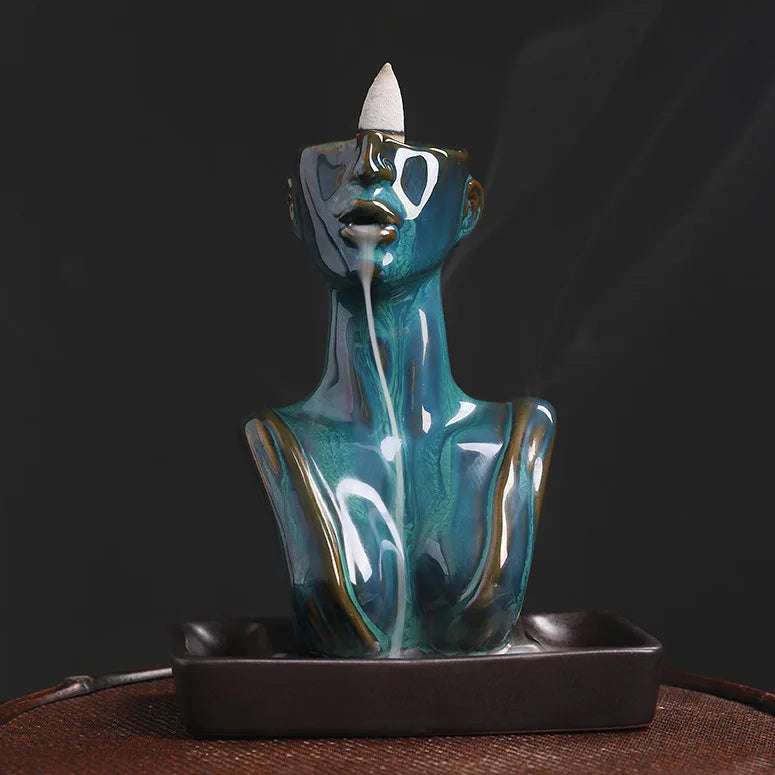 Full front view of a blue abstract human bust backflow incense burner on a woven mat, demonstrating the smoke waterfall effect flowing from the mouth to the base tray.