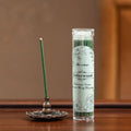 Glow & Drift cedarwood incense sticks in a clear tube with a green stick burning in a bronze lotus incense holder.
