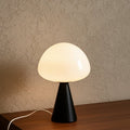 Glow & Drift Eclipse mushroom table lamp with a matte black iron base and glowing white glass dome on a wooden surface.