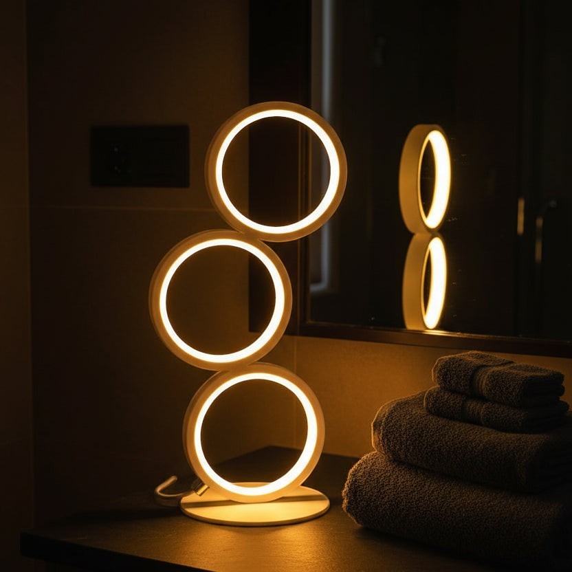 Glow & Drift Eclipse Triple Ring Table Lamp with warm LED illumination on a dark surface next to folded towels.