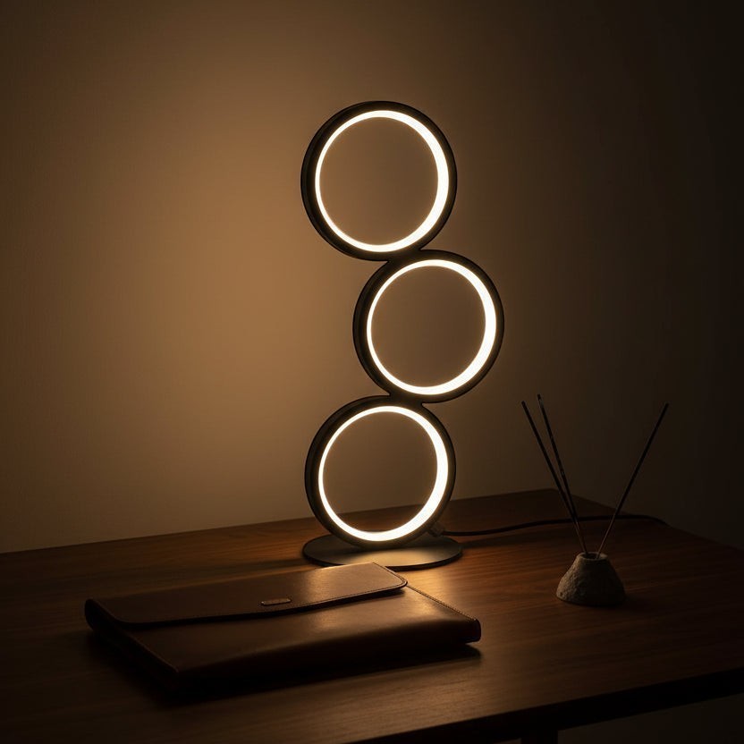 Glow & Drift Eclipse Triple Ring Table Lamp with modern black sculptural design emitting warm ambient light on a desk.