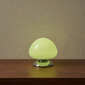 Glow & Drift Aura glass mushroom table lamp with a glowing light green dome and polished chrome base on a wood table.
