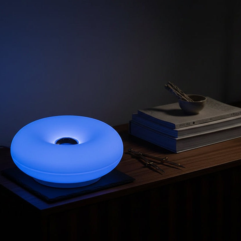 Glow & Drift Halo Donut Spectrum portable lamp glowing with blue light on a wooden bedside table with books.