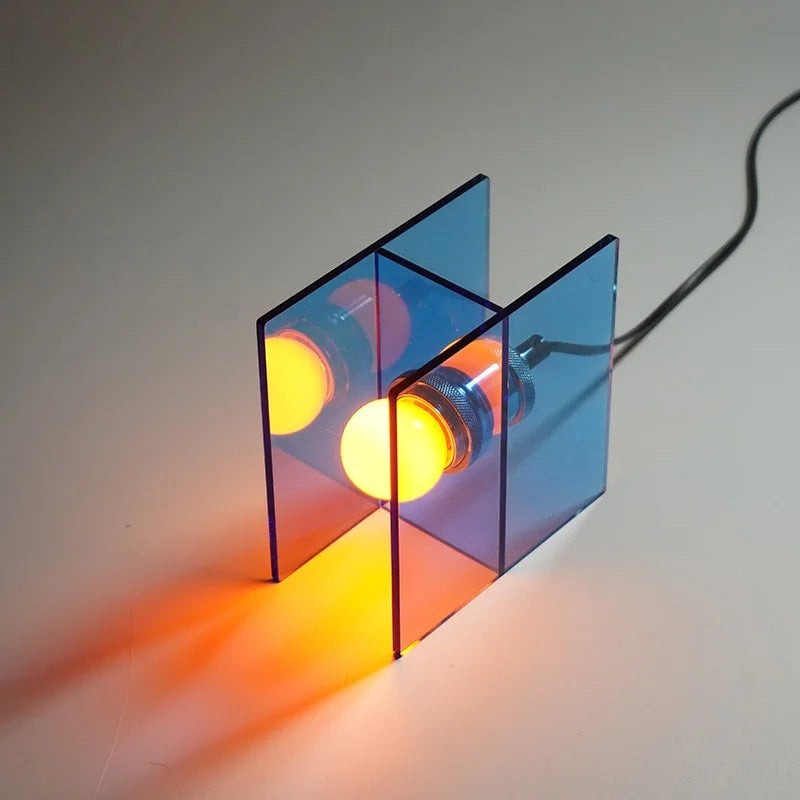 Glow & Drift Horizon geometric table lamp with blue acrylic panels and a warm glowing light bulb on a white surface.