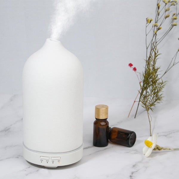 A Chalk White frosted ceramic humidifier by Glow and Drift actively diffusing a soothing, cool mist, sitting on a clean white marble countertop alongside small amber oil bottles.