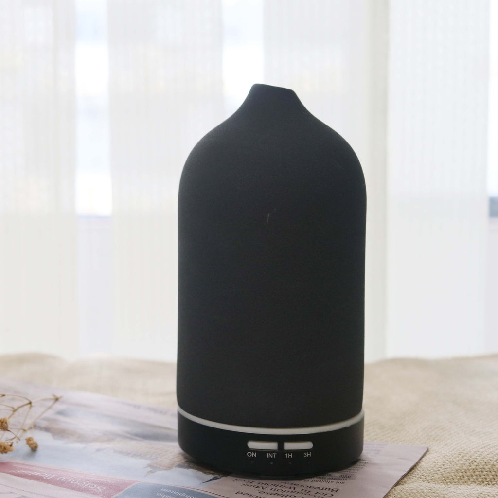 A Charcoal Black textured ceramic ultrasonic diffuser by Glow and Drift, resting elegantly on a folded newspaper with a bright, airy window in the background.