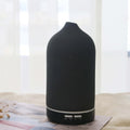 A Charcoal Black textured ceramic ultrasonic diffuser by Glow and Drift, resting elegantly on a folded newspaper with a bright, airy window in the background.