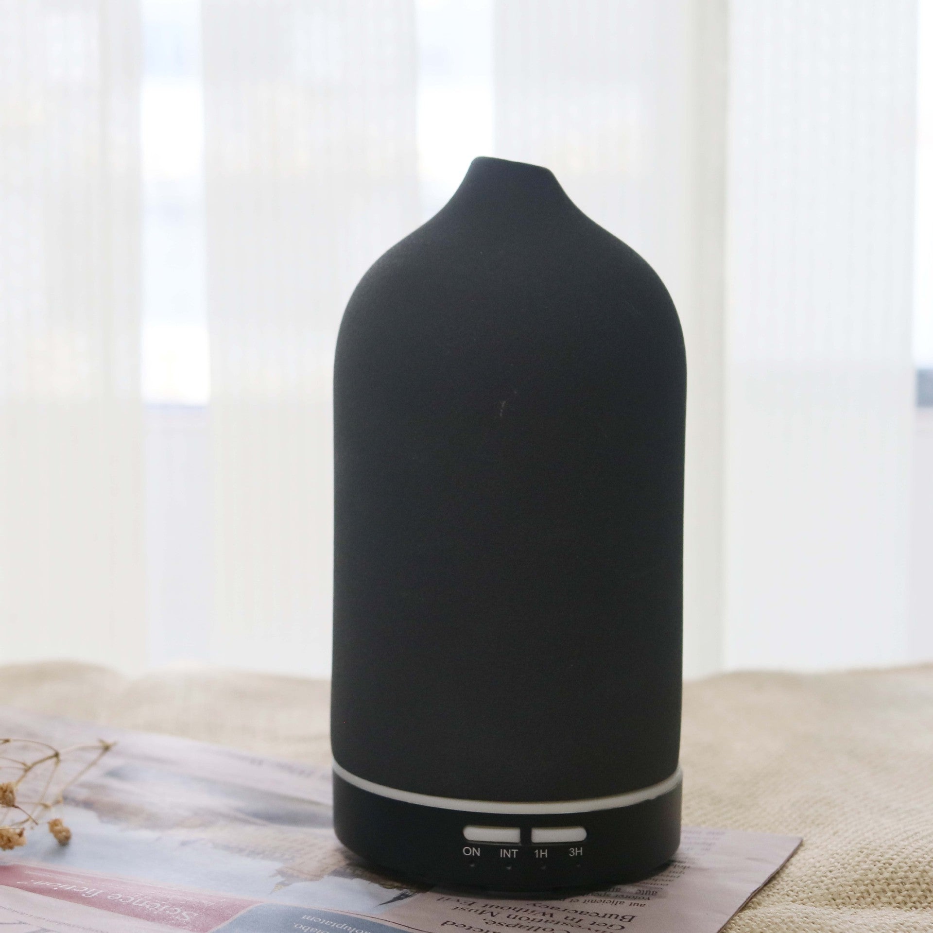 A Charcoal Black textured ceramic ultrasonic diffuser by Glow and Drift, resting elegantly on a folded newspaper with a bright, airy window in the background.