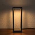Glow & Drift Nomad Lantern, a minimalist black geometric cordless lamp casting a warm, soft glow on a wooden surface.