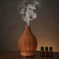 Glow & Drift Artisan Woven Vase Diffuser with rattan-style shell emitting mist next to three essential oil bottles.