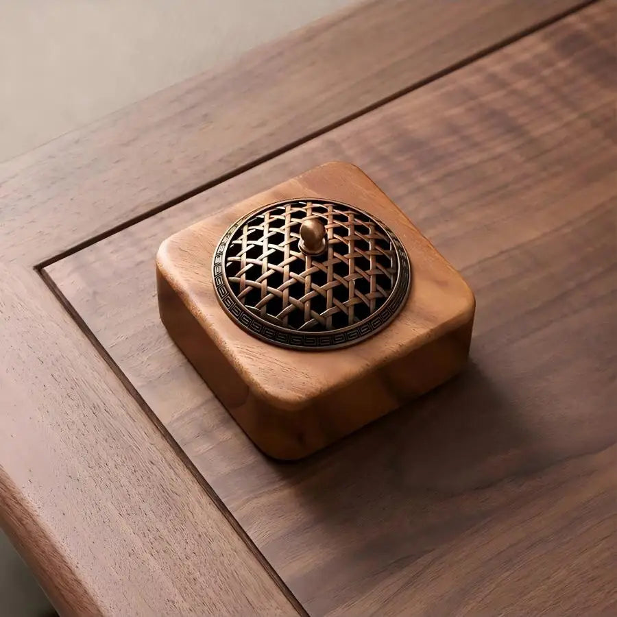 Square light wood aromatherapy incense burner featuring a woven brass metal lid resting on a rich walnut desk.