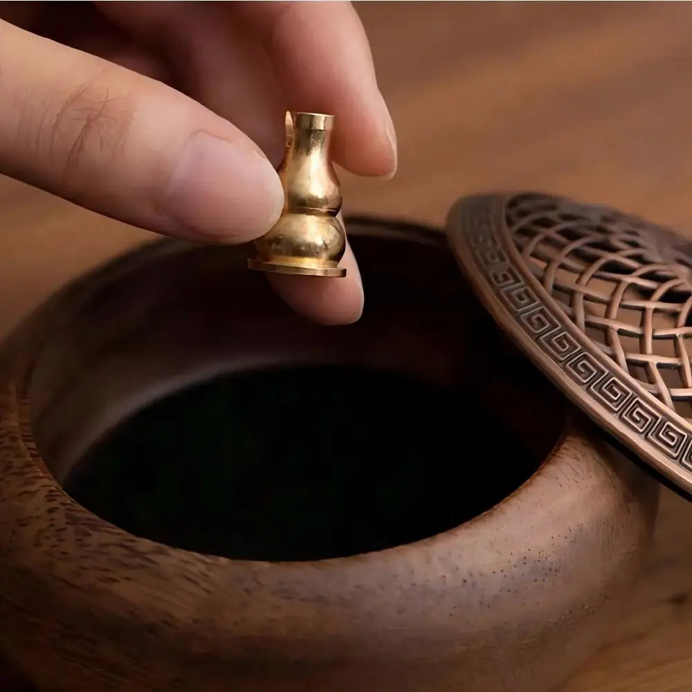 A hand delicately holding the mini brass gourd-shaped incense accessory over the open wooden base of the aromatherapy furnace.