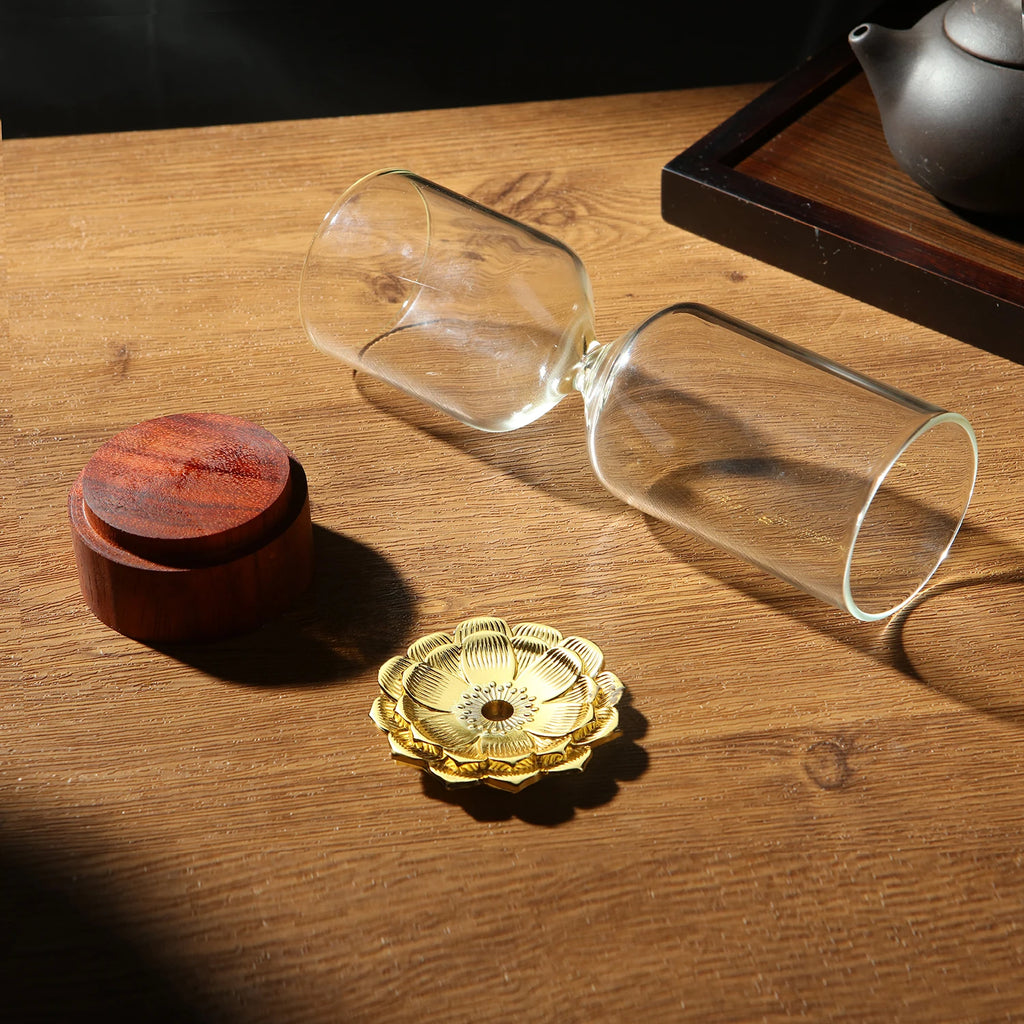 The disassembled parts of The Hourglass Waterfall Incense Vessel laid out on a wooden table, displaying the dark wood base, the clear glass hourglass body, and the intricate gold lotus incense cone holder.