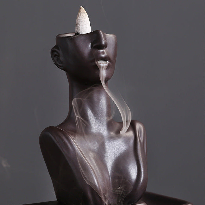 Angled close-up of a matte obsidian backflow incense burner shaped like a human bust, showcasing a thick, mesmerizing stream of smoke flowing gracefully from the lips and curling down the torso.