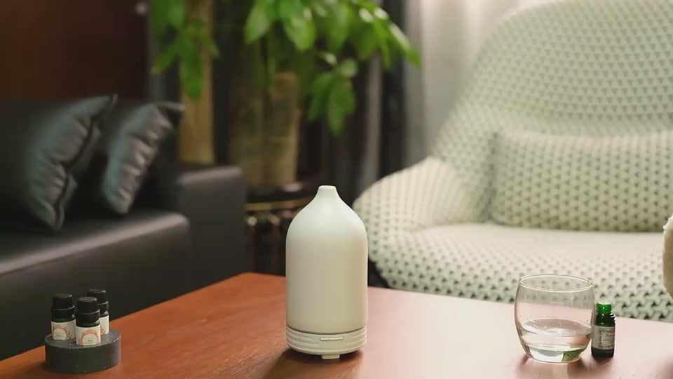 A product demonstration video showing a person opening the Minimalist Frosted Ceramic Diffuser, lifting the matte white outer shell and inner lid, pouring water from a glass tumbler into the 100ml reservoir, adding drops of essential oil, replacing the covers, and turning on the power button to emit a smooth, soothing stream of ultrasonic aroma mist on a wooden coffee table.