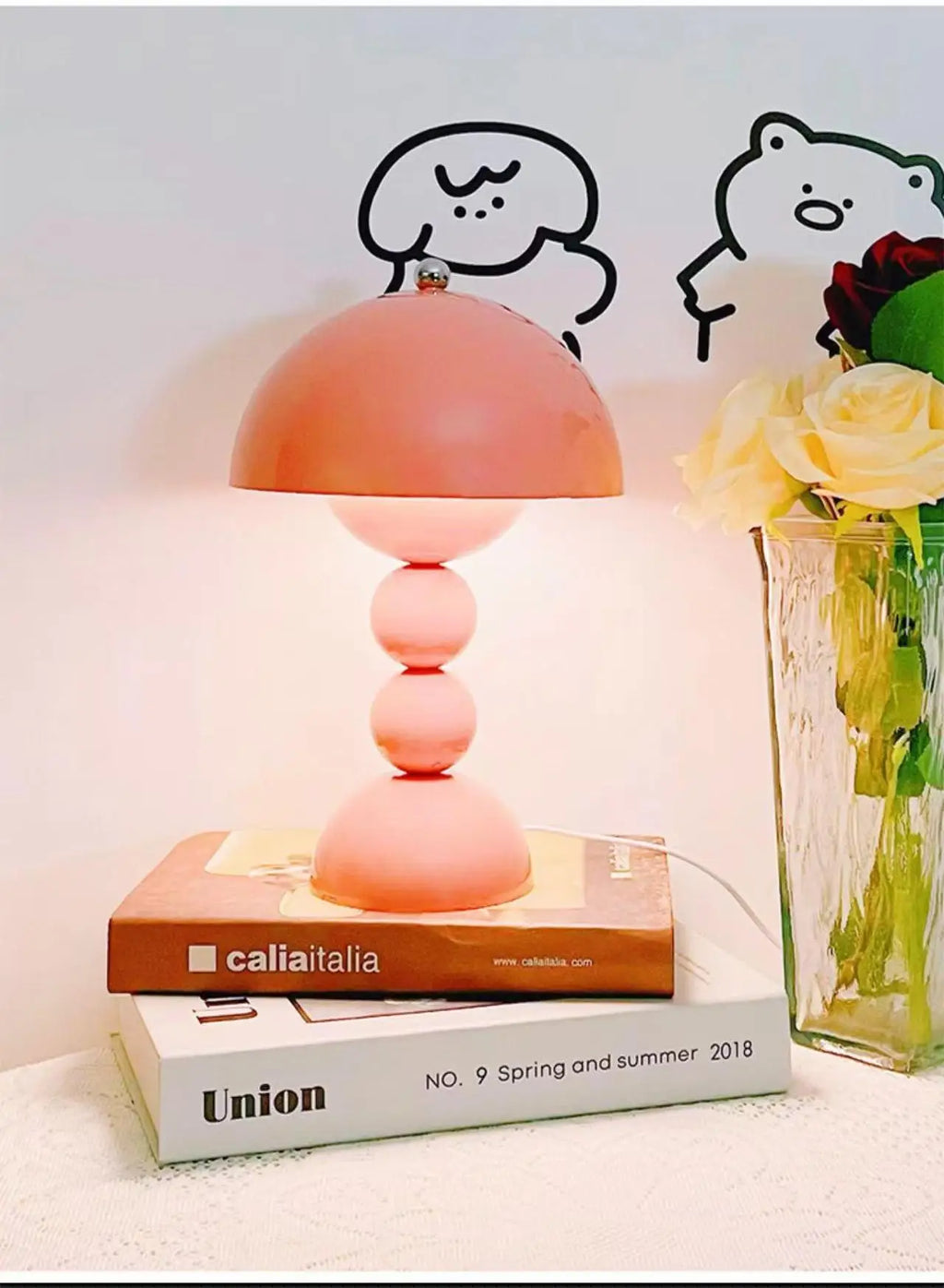 A soft pink Totem creative table lamp resting on stacked decorative books next to a textured glass vase with white roses.