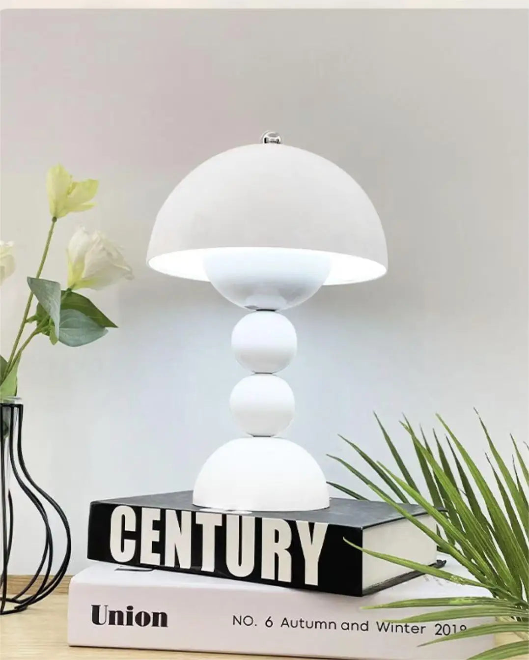 A clean white Totem modern table lamp styled on top of display books next to a vase with delicate white flowers and green palm leaves.