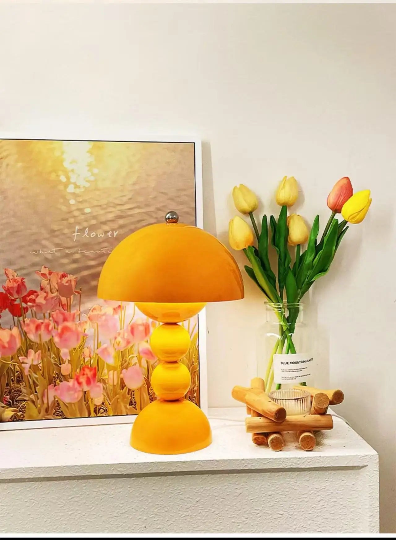 A bright yellow Totem Nordic LED table lamp styled on a white shelf surrounded by fresh tulips and a floral art piece.