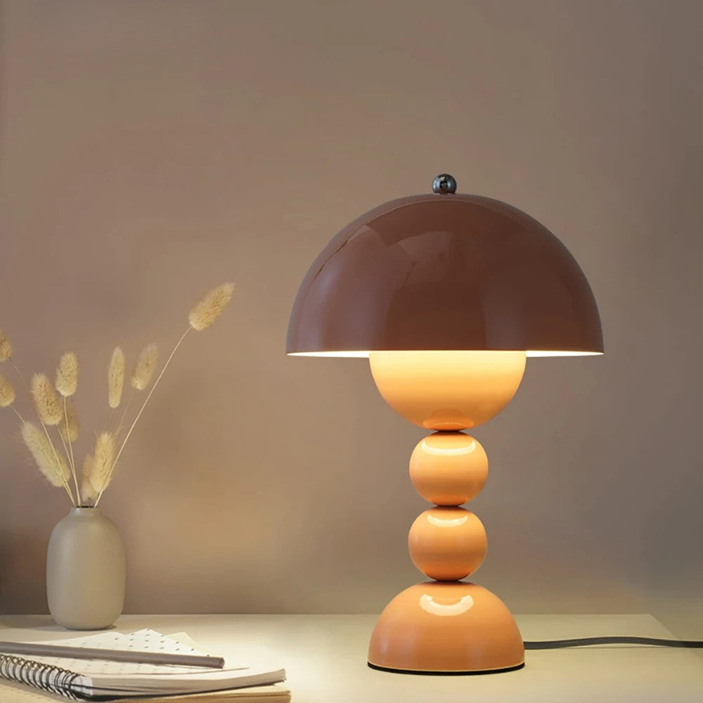 A tan-colored Totem LED desk lamp illuminated on a wooden workspace next to a notebook, pen, and dried bunny tail grass in a vase.