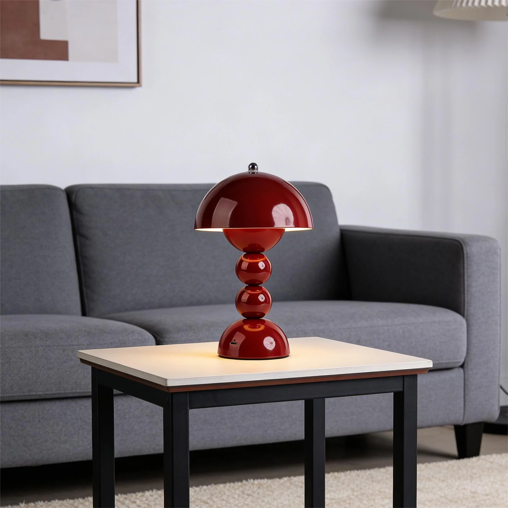 A glossy red Totem rechargeable LED lamp illuminating a white-topped side table in front of a cozy grey fabric sofa in a minimalist living room.