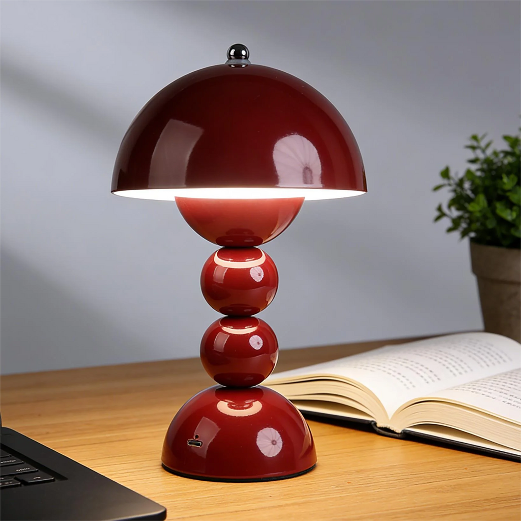 A close-up of the deep red Totem stepless dimming desk lamp glowing over an open book on a clean wooden table next to a small potted plant.